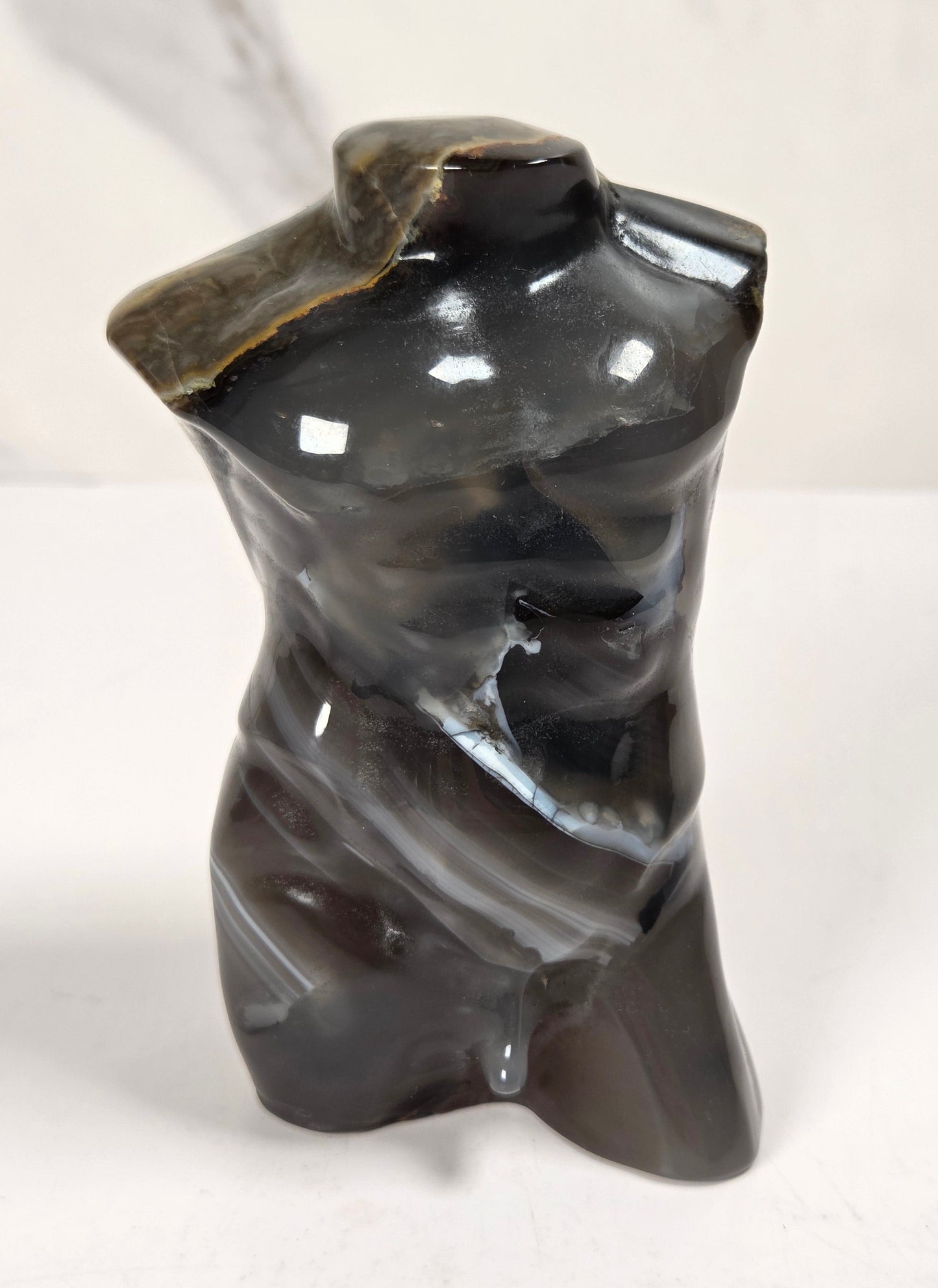 A small glossy black stone torso sculpture with natural striated texture and polished highlights, photographed on a light surface against a pale marble-look background.