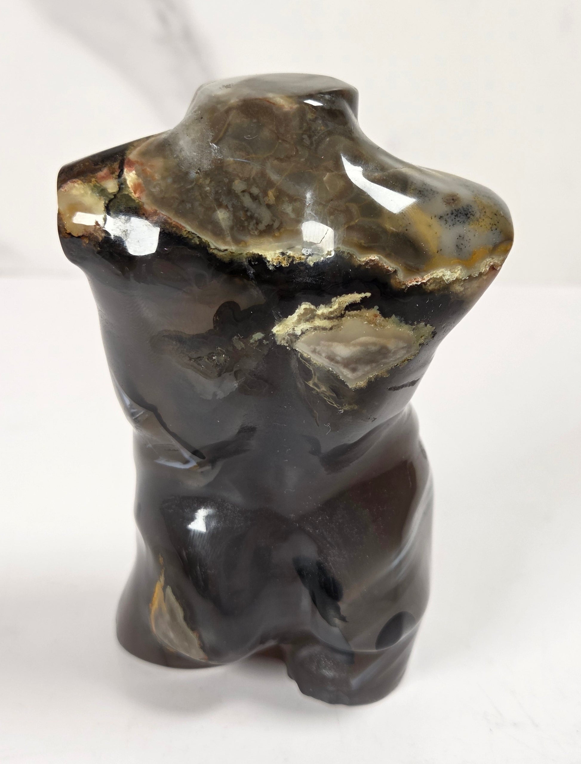 A small polished stone sculpture shaped like a stylized torso, shown on a white surface with a light marble-patterned background.