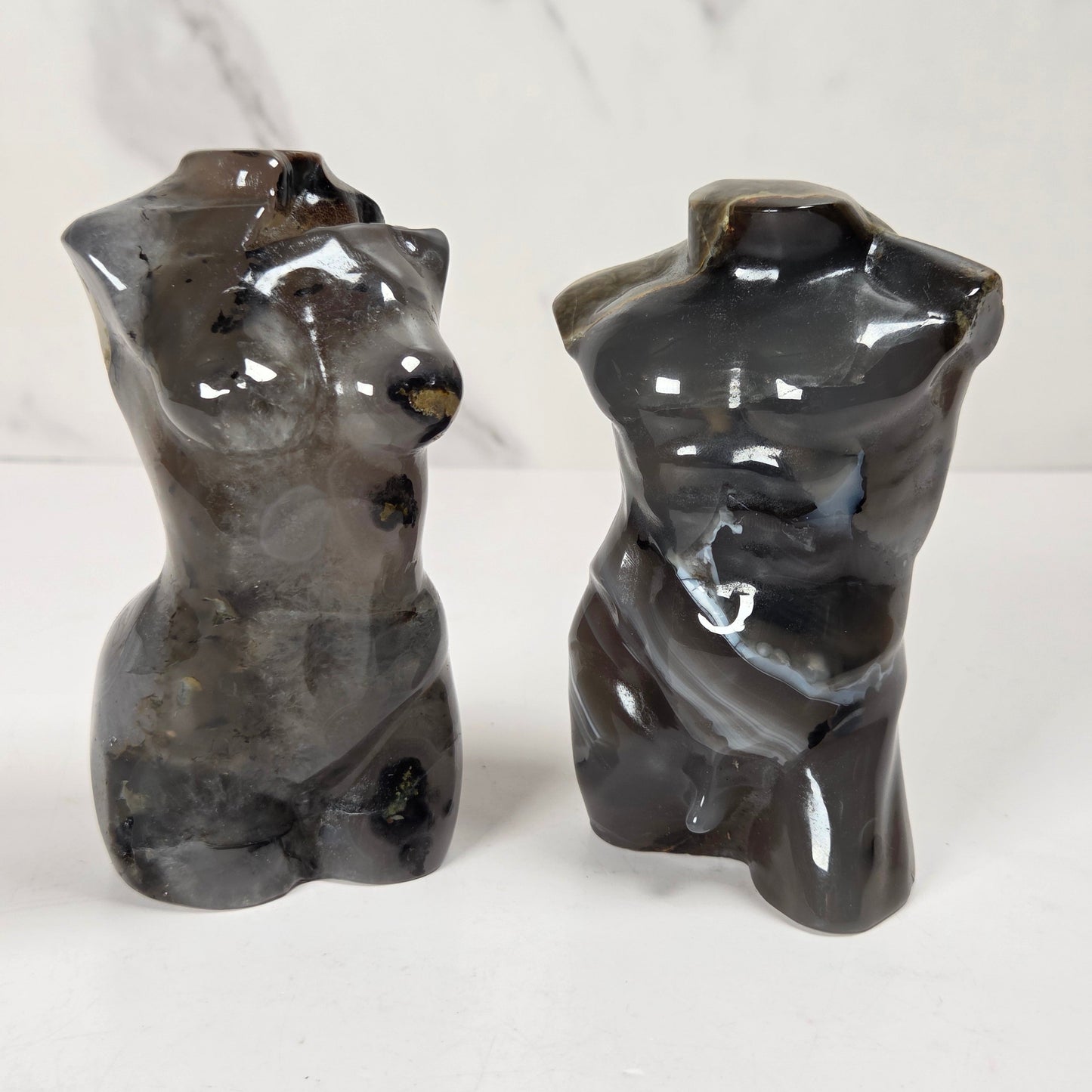 Two glossy abstract torso sculptures in dark gray stone, shown side by side on a white surface against a light marble-patterned background.