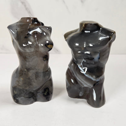 Two glossy abstract torso sculptures in dark gray stone, shown side by side on a white surface against a light marble-patterned background.