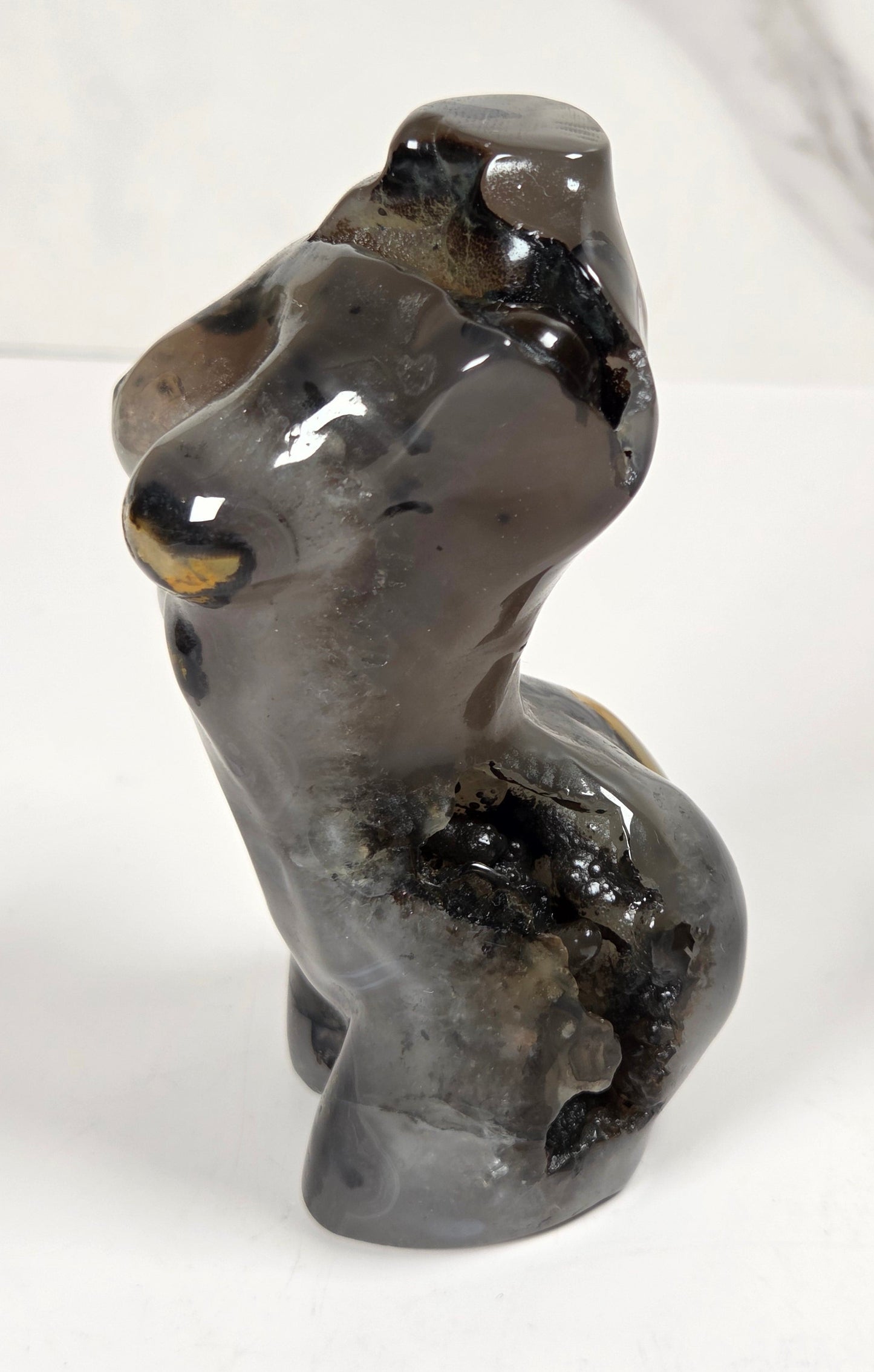 A small glossy stone figurine of a seated figure, likely carved from dark gray to black stone with subtle warm brown and gold highlights, set against a light gray marble-patterned background.