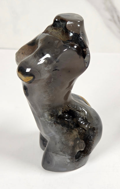 A small glossy stone figurine of a seated figure, likely carved from dark gray to black stone with subtle warm brown and gold highlights, set against a light gray marble-patterned background.