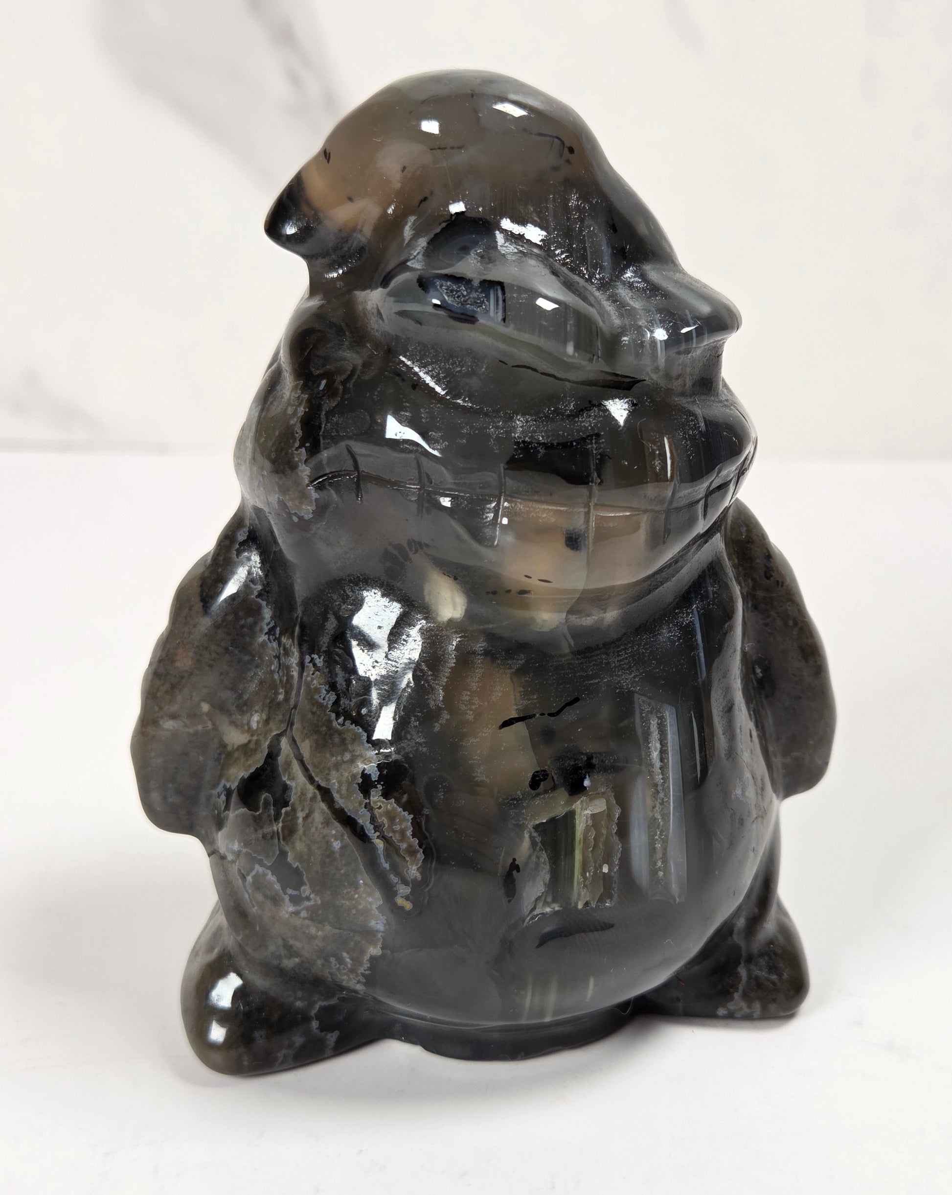 A small glossy carved figurine of a frog or toad made from smoky translucent stone, standing on a white surface against a light marble-look background.