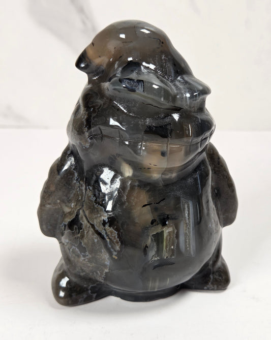 A small glossy carved figurine of a frog or toad made from smoky translucent stone, standing on a white surface against a light marble-look background.