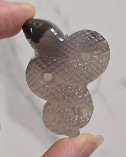 A hand holding a carved stone pendant shaped like a snake, featuring a textured diamond pattern and a dark polished head, set against a light marble background.