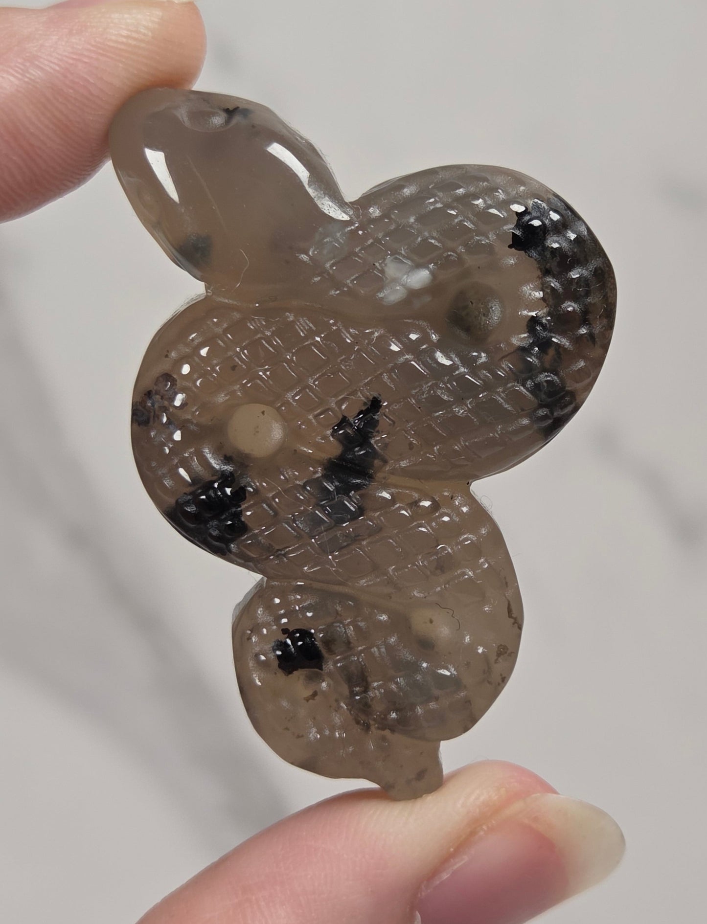 A hand holding a carved snake-shaped pendant or charm made of translucent gray stone with black inclusions and a textured pattern.