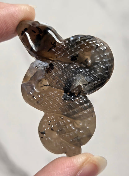 A hand holding a carved snake pendant made of translucent smoky brown and black stone, shown against a light marble-like background.