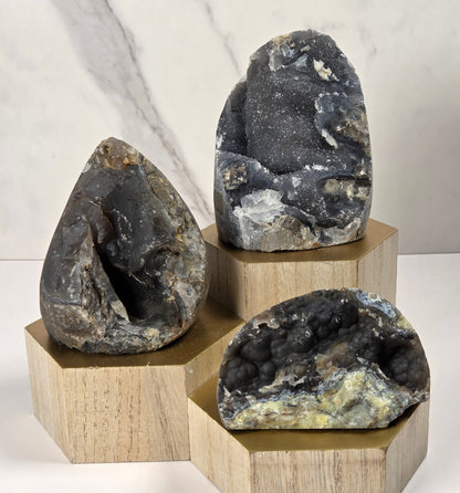 Three natural geode specimens with dark, crystalline interiors displayed on light wood hexagonal pedestals against a white and gray marble-patterned background.