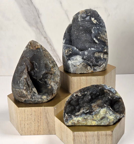 Three natural geode specimens with dark, crystalline interiors displayed on light wood hexagonal pedestals against a white and gray marble-patterned background.