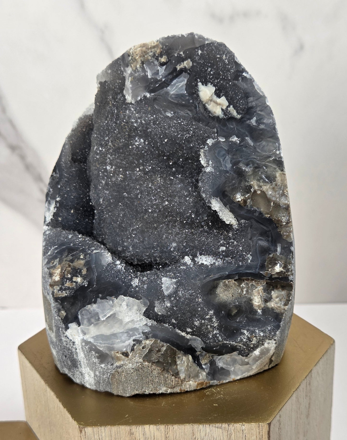 A dark gray geode specimen with white and light gray crystalline formations, displayed on a hexagonal gold-colored wooden pedestal against a white marble-patterned background.