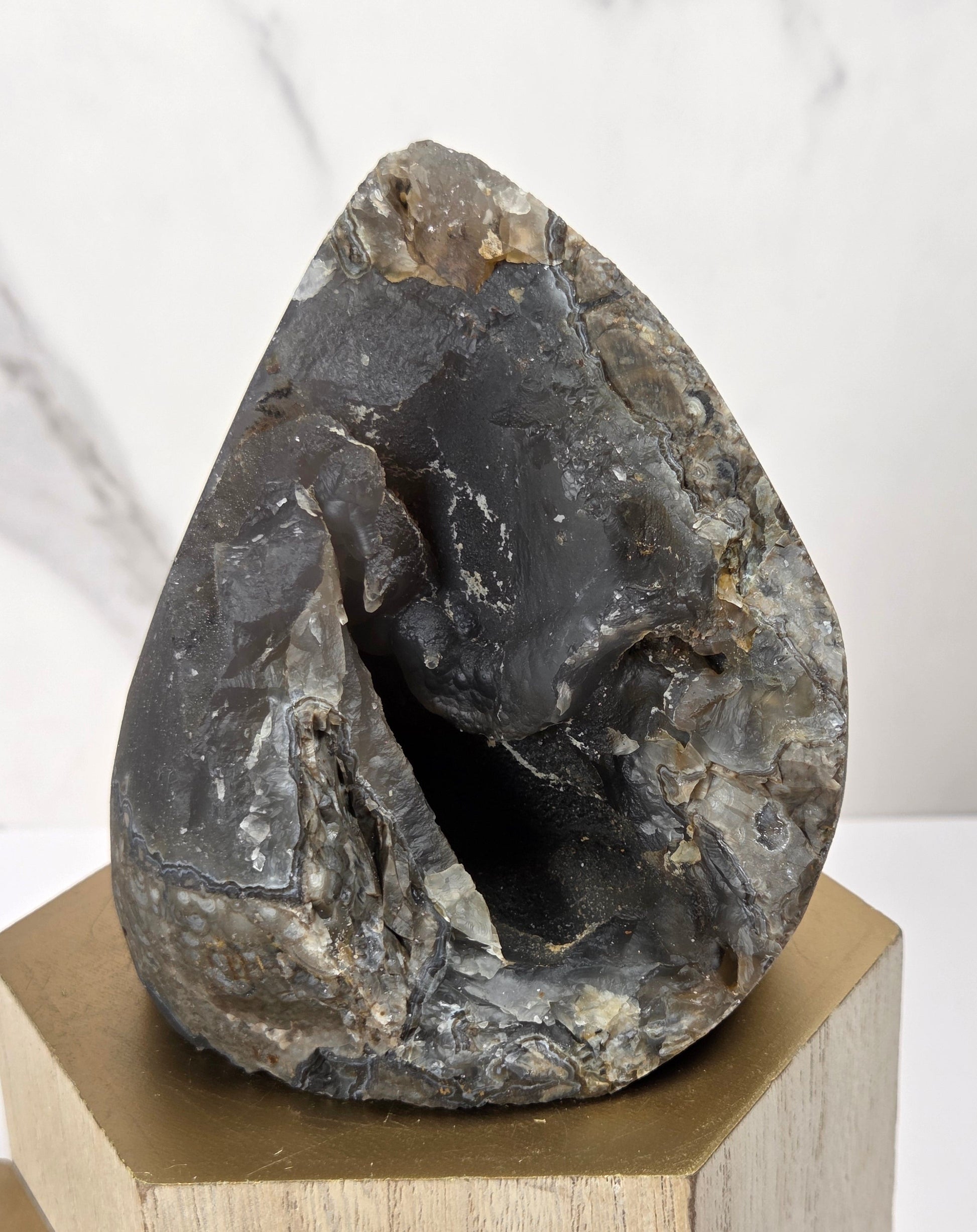 A raw, polished dark gray stone with a hollow cavity, displayed on a light wood block pedestal against a white marble background.