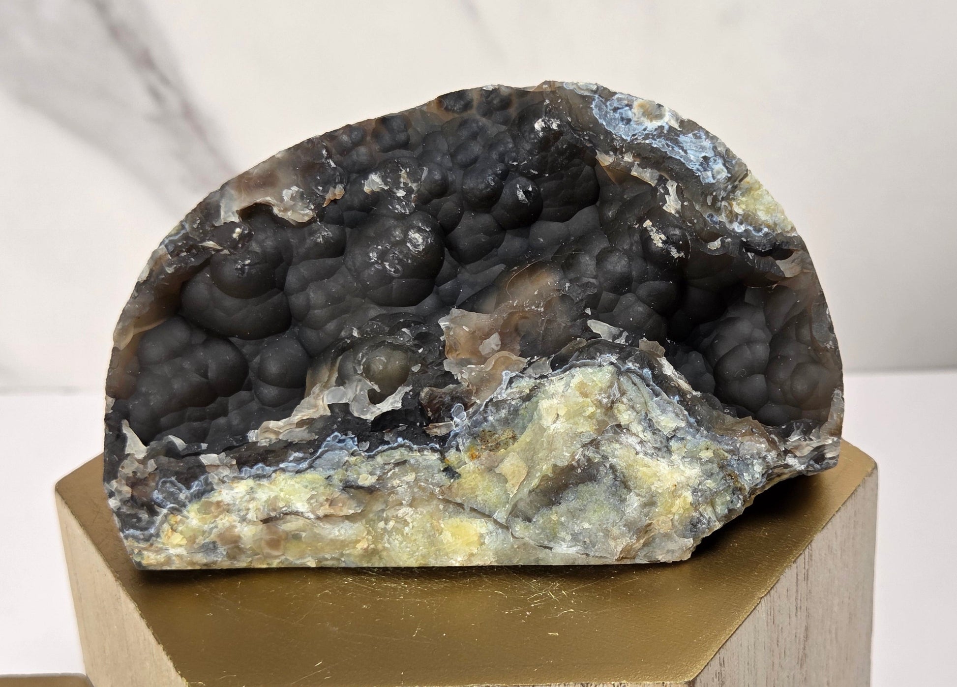 A natural geode specimen with a dark, bubble-like interior and pale yellow to blue-gray outer layers, displayed on a gold-toned geometric pedestal against a light marble-patterned background.