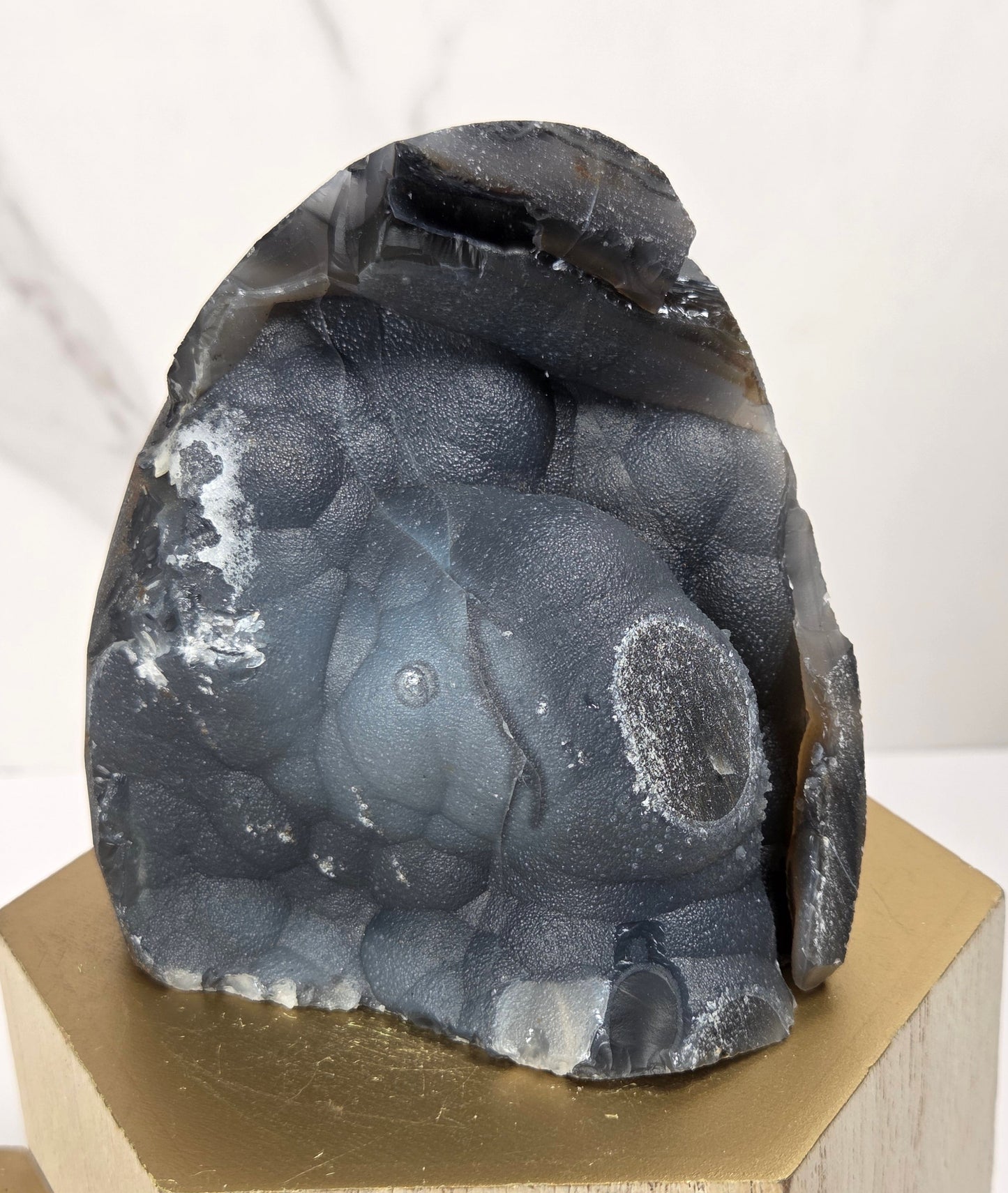 A dark gray and black textured stone sculpture displayed on a light wood hexagonal pedestal against a white marble-patterned background.
