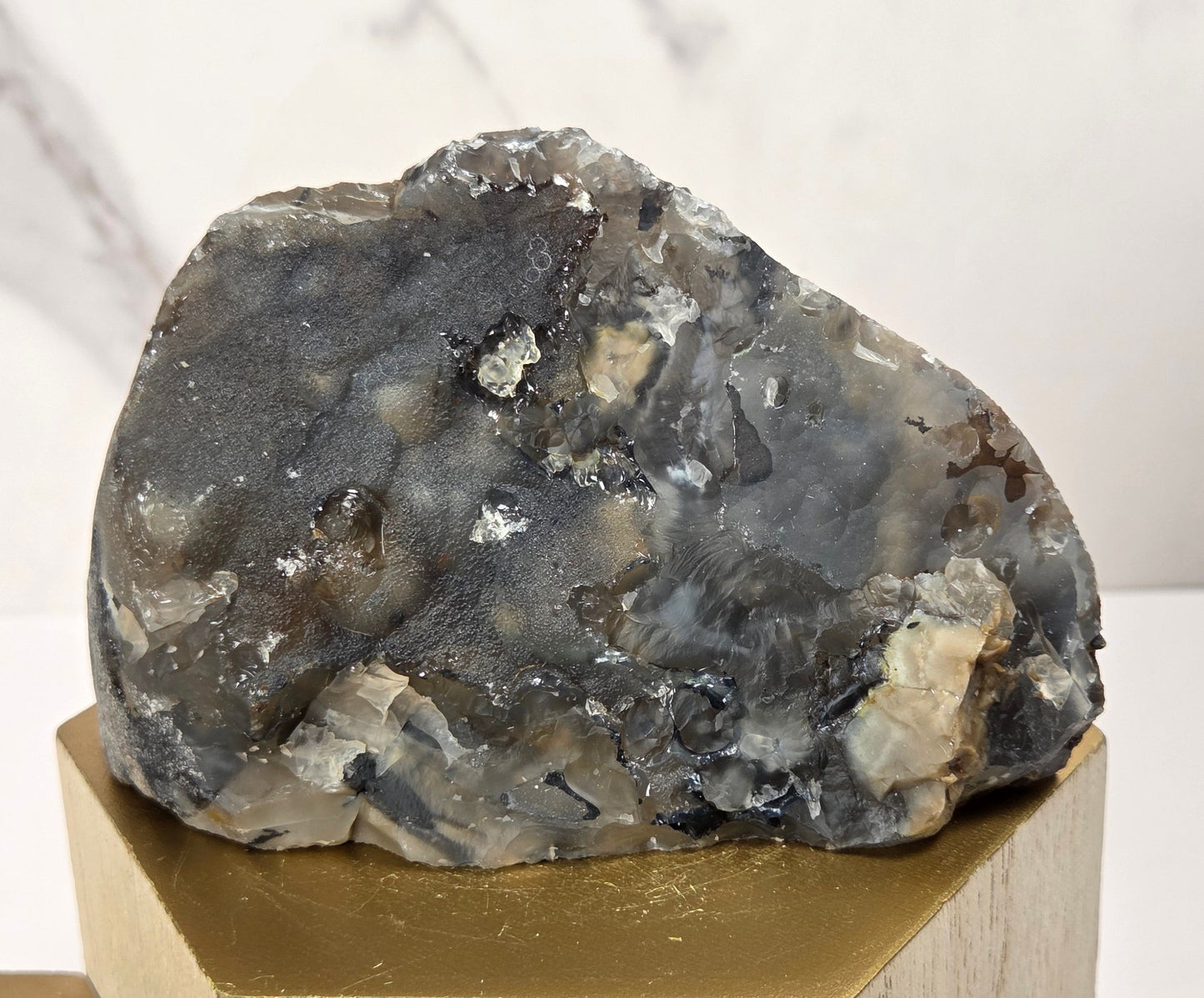 A rough natural stone or mineral specimen with a mottled gray, black, and off-white surface, displayed on a hexagonal gold-toned pedestal against a light marble-patterned background.