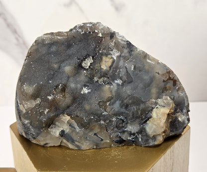 A rough natural stone or mineral specimen with a mottled gray, black, and off-white surface, displayed on a hexagonal gold-toned pedestal against a light marble-patterned background.