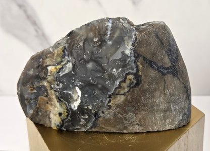 A raw agate crystal specimen with layered gray, black, and brown bands, displayed on a gold hexagonal pedestal against a white marble background.