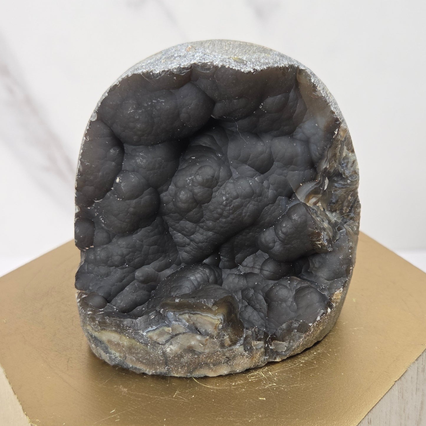 A dark gray geode specimen with a textured, bubbly interior, displayed on a gold hexagonal pedestal over a light wood block against a white marble background.