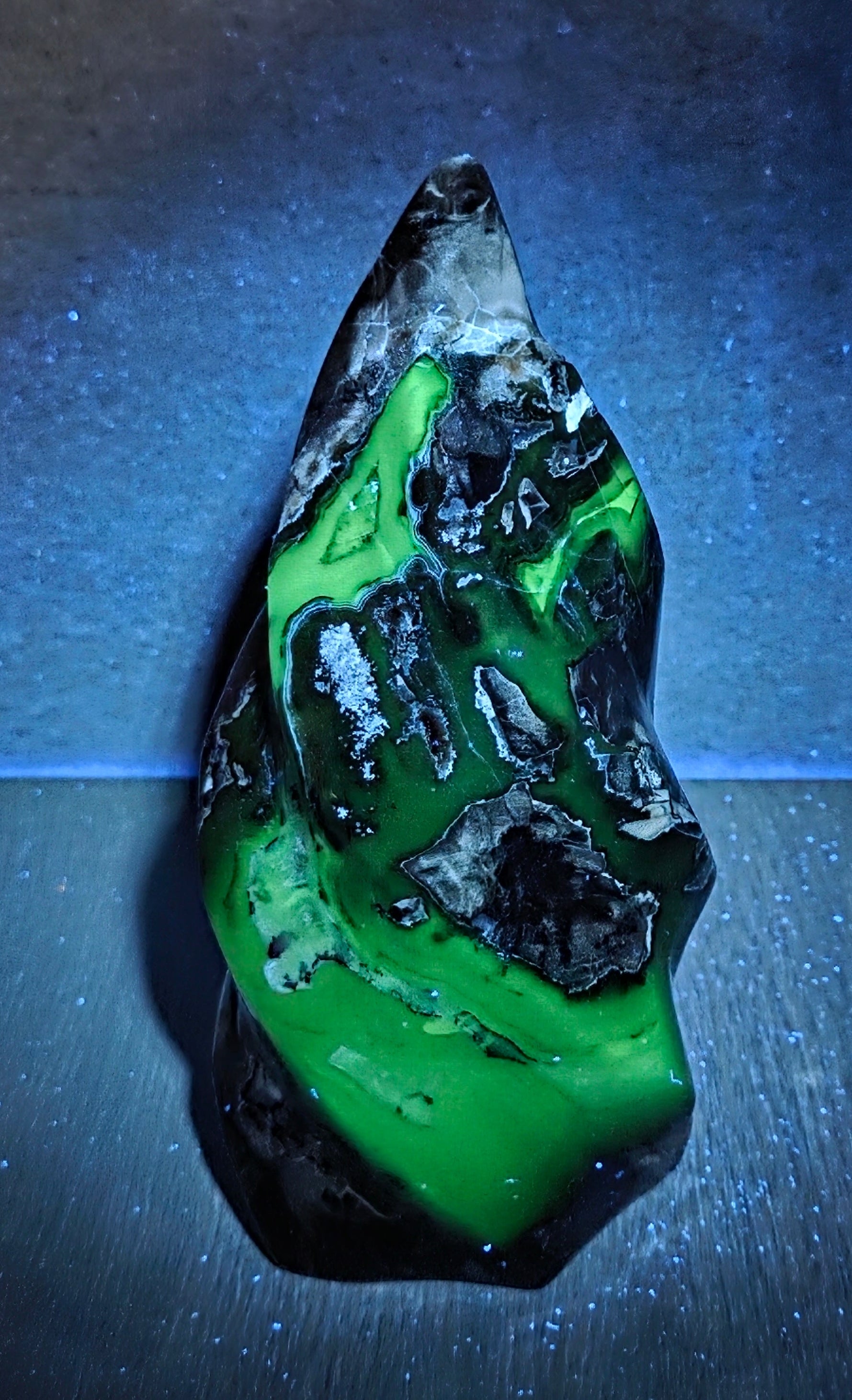 A polished green and black mineral specimen or raw gemstone sculpture standing upright on a dark surface under blue spotlighting.