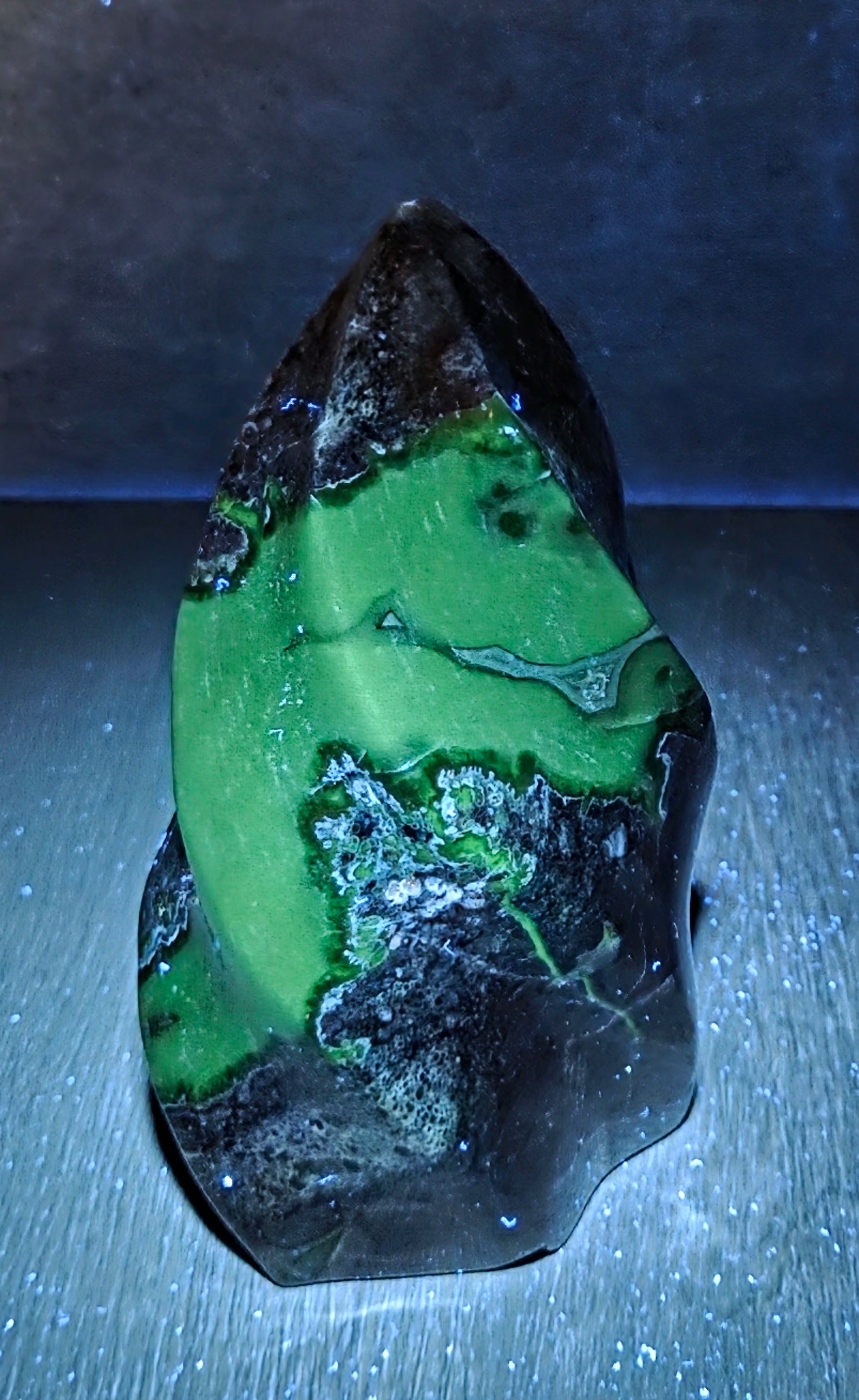 A polished natural stone carving with a pointed top, featuring a vibrant green body with prominent black and dark gray veining, displayed on a dark textured surface under dramatic blue lighting.