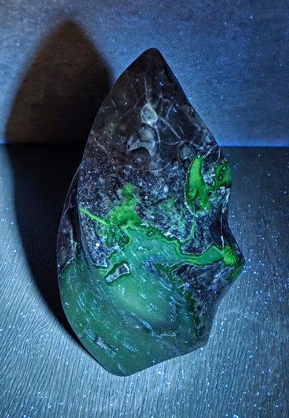 A raw, polished mineral specimen with vibrant green and black mottled patterns, displayed on a dark surface under blue light.