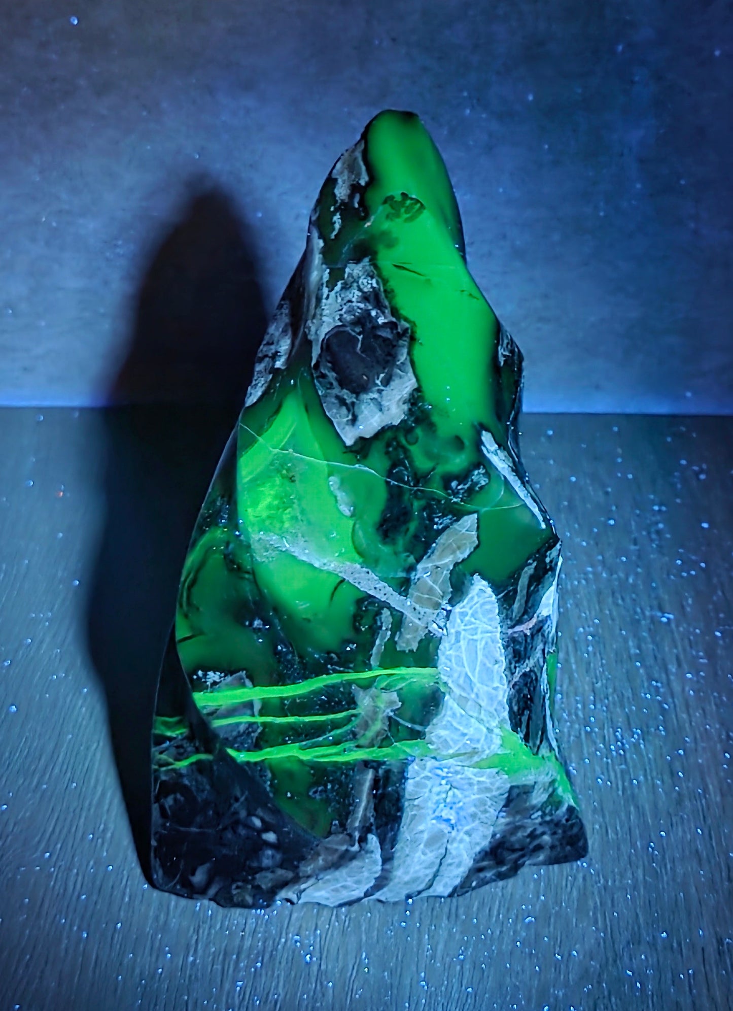 A raw, pointed mineral specimen with vibrant neon green and black matrix, displayed on a dark surface under blue light.