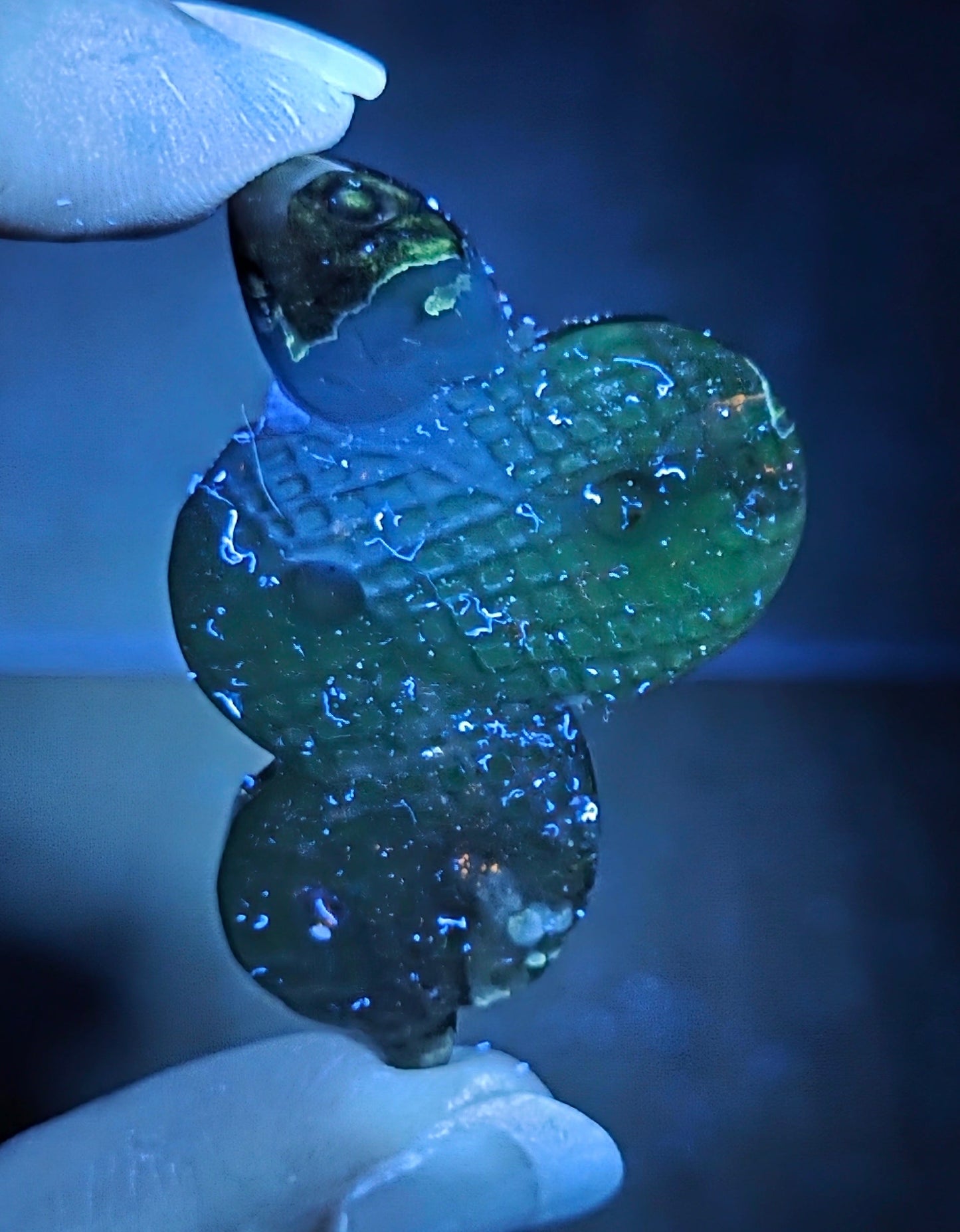 A hand holding a small, three-blobed translucent green glass bead pendant under blue light, showing a textured pattern.