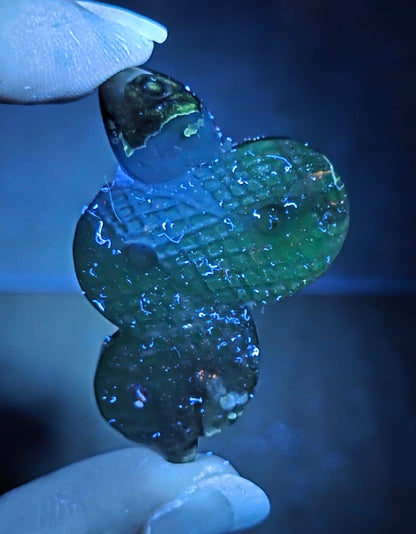 A hand holding a small, three-blobed translucent green glass bead pendant under blue light, showing a textured pattern.