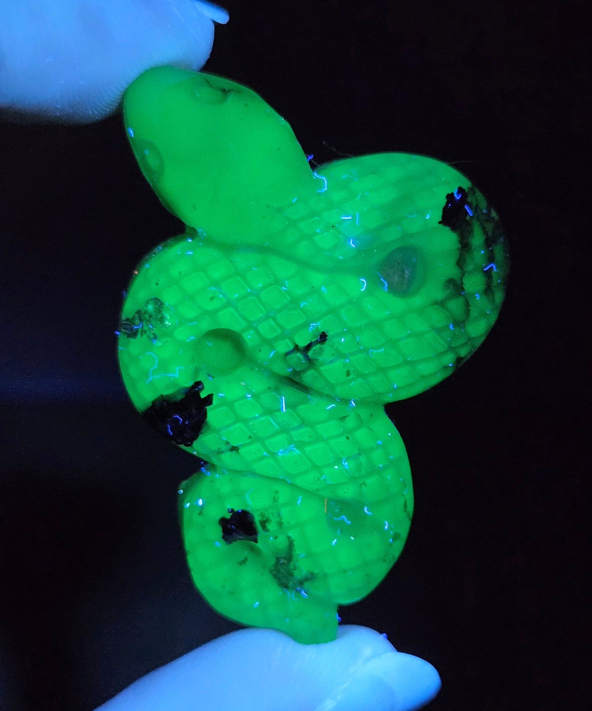 A small coiled green snake-shaped pendant or charm with a textured scale pattern, held between two gloved fingers against a very dark background.
