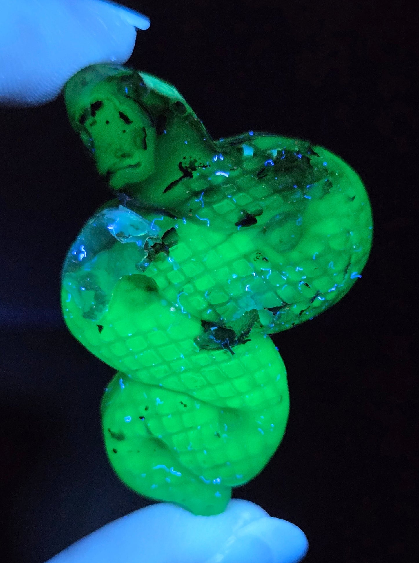A small coiled pendant or charm made of translucent green material, held between gloved fingers against a very dark background.
