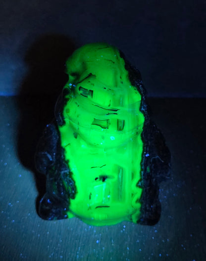 A small glowing neon green figurine or decorative object with dark sides, photographed against a very dark background that fluoresces blue-green under UV light.