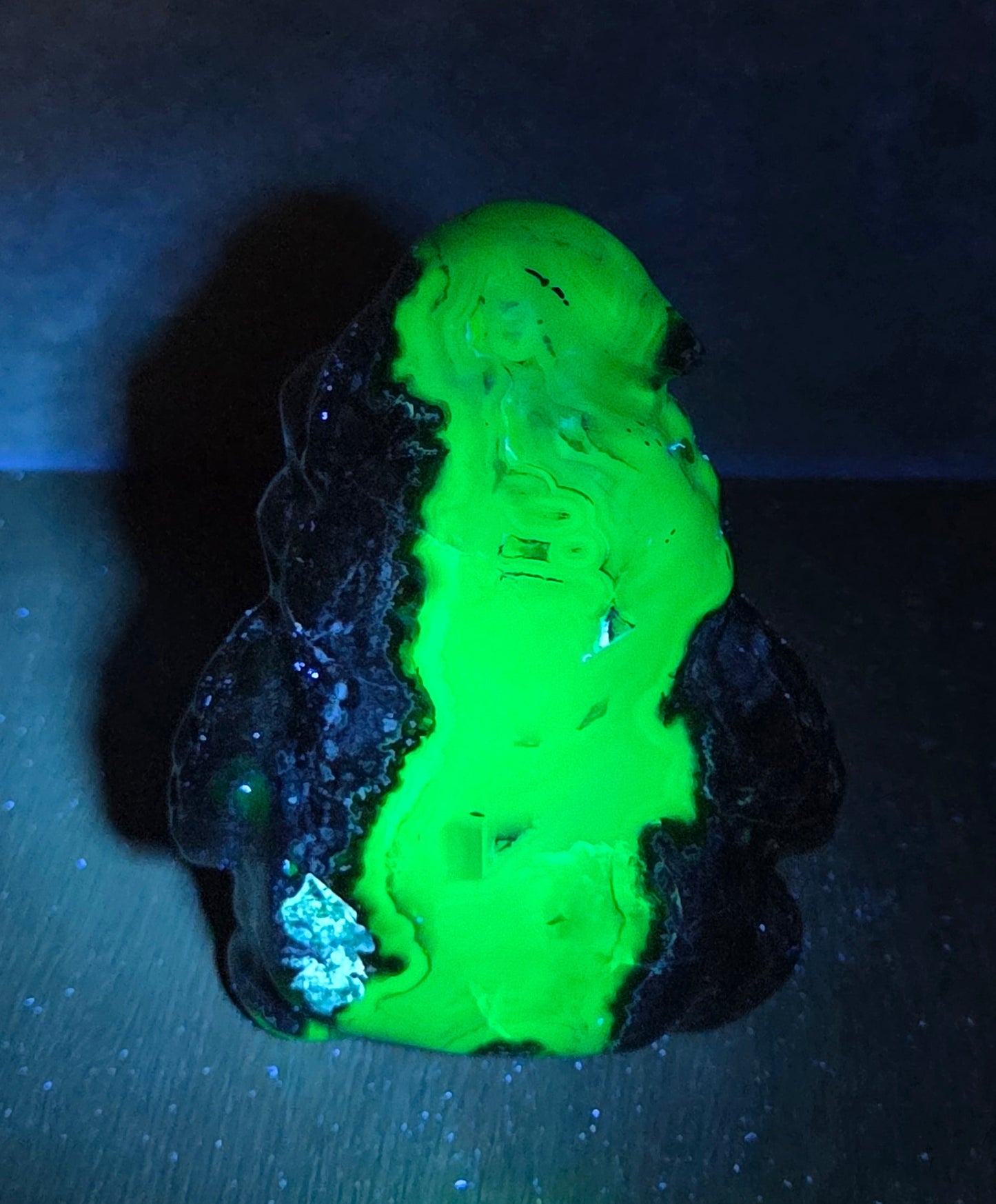 A small novelty figurine or decorative object with a bright fluorescent green front and dark, textured sides, photographed on a dark surface against a dark background under blue-violet light.
