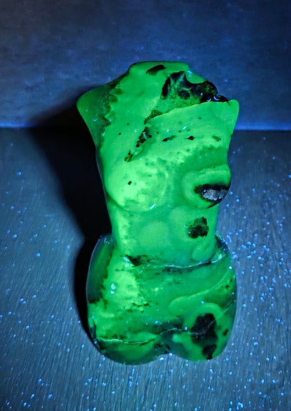 A small sculpted bust figure glowing bright green under UV light, placed on a dark surface with a blue-lit background.