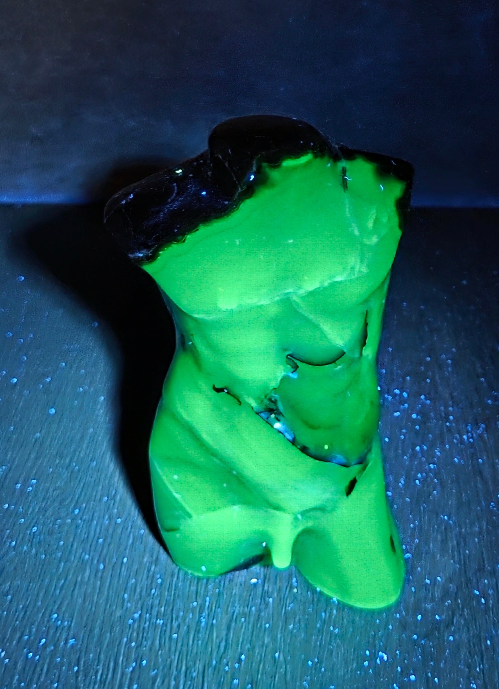 A small green figurine or sculpture with black hair, photographed on a dark surface under blue light. The product appears to be the figurine itself rather than packaging or an instructional graphic.