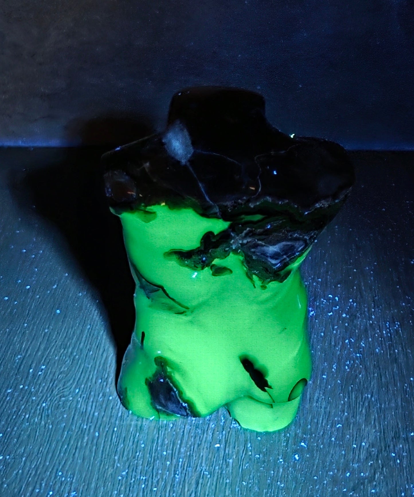 A small sculptural object with a bright neon green body and a dark, rough black top, photographed on a dark surface against a dark background under blue light.