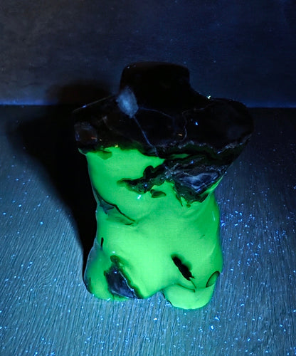 A small sculptural object with a bright neon green body and a dark, rough black top, photographed on a dark surface against a dark background under blue light.