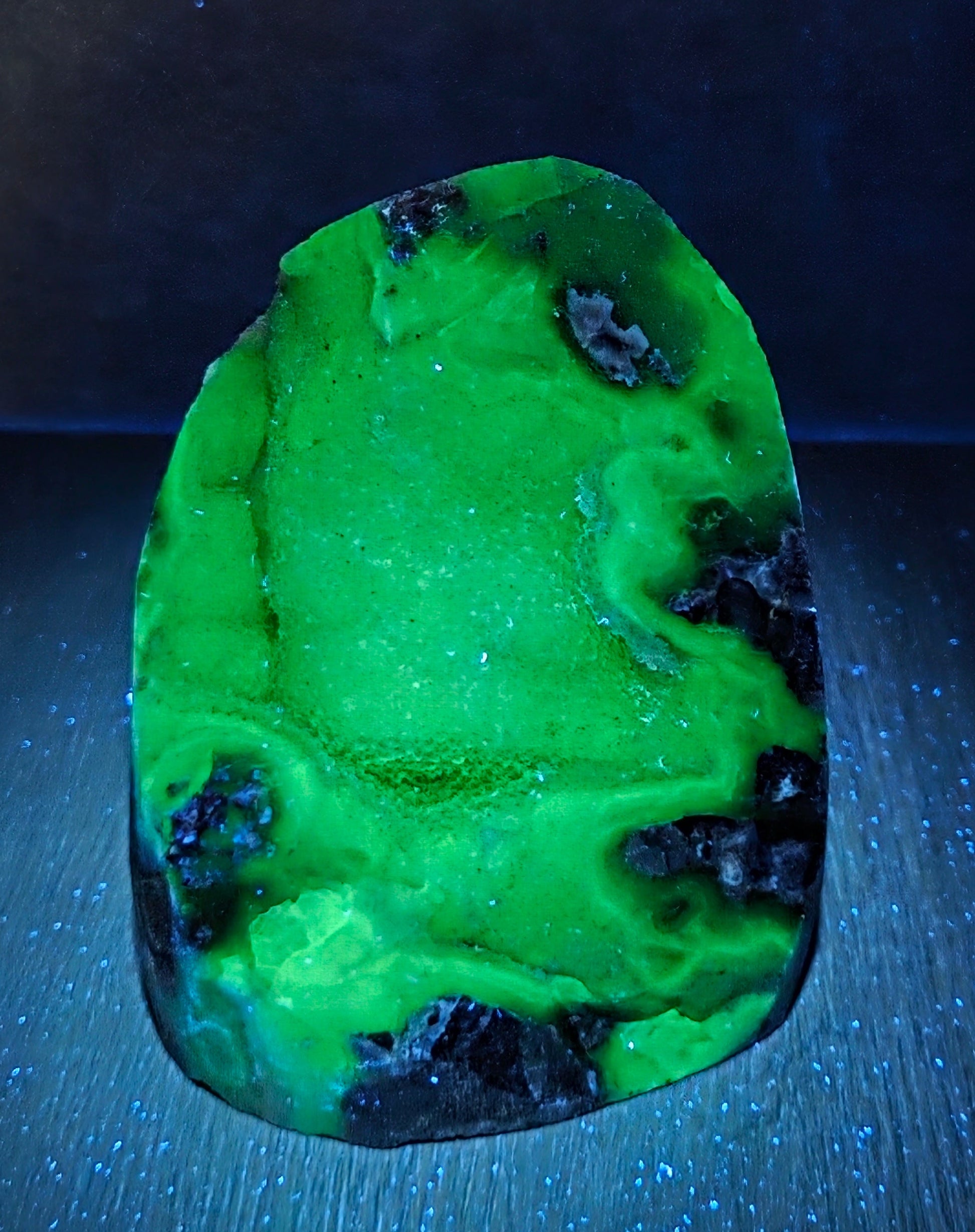 A raw, polished green stone or mineral specimen with black inclusions, displayed on a dark textured surface under blue light.
