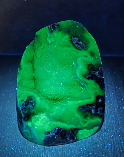 A raw, polished green stone or mineral specimen with black inclusions, displayed on a dark textured surface under blue light.