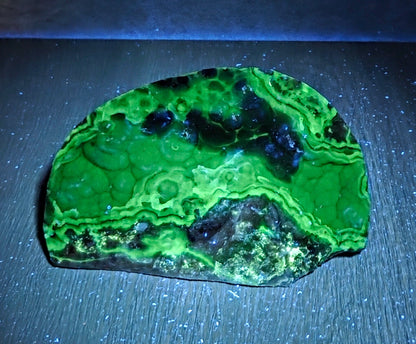 A raw green malachite mineral specimen with dark blue-black inclusions, displayed on a dark textured surface under blue light.