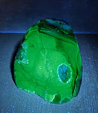 A raw green stone or mineral specimen with natural marbling and a circular inclusion, photographed on a dark surface under blue light.