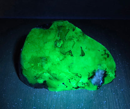 A glowing green mineral specimen or raw gemstone placed on a dark surface under blue-violet light, with a black background.