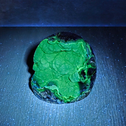 A small irregularly shaped stone or mineral sample with a bright neon green marbled surface and dark edges, photographed on a dark blue-lit surface.