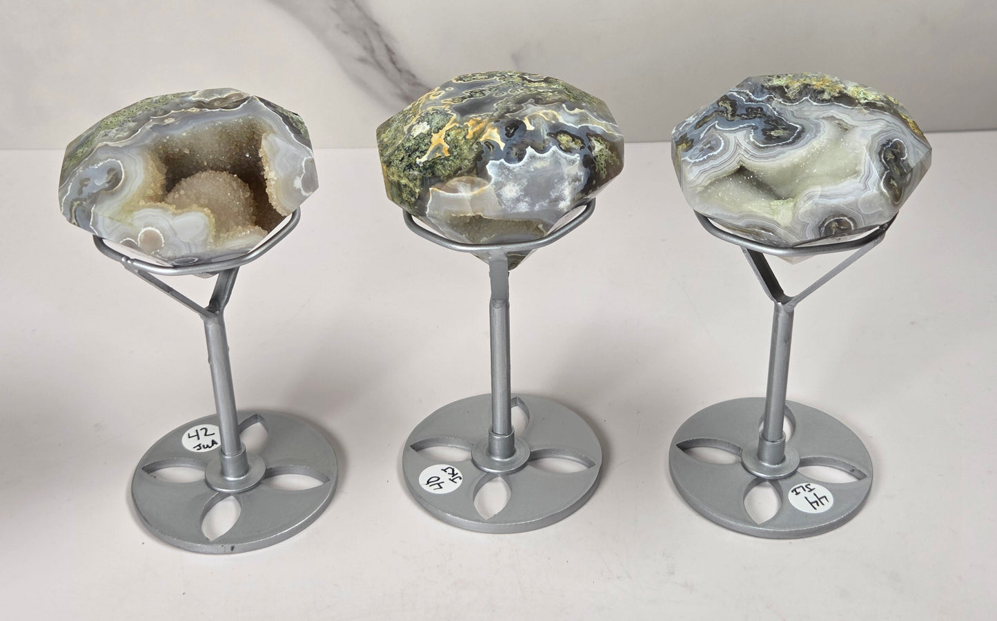 Three polished agate geode specimens displayed on silver metal stands against a light gray marble-patterned background.
