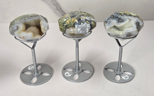 Three polished agate geode specimens displayed on silver metal stands against a light gray marble-patterned background.
