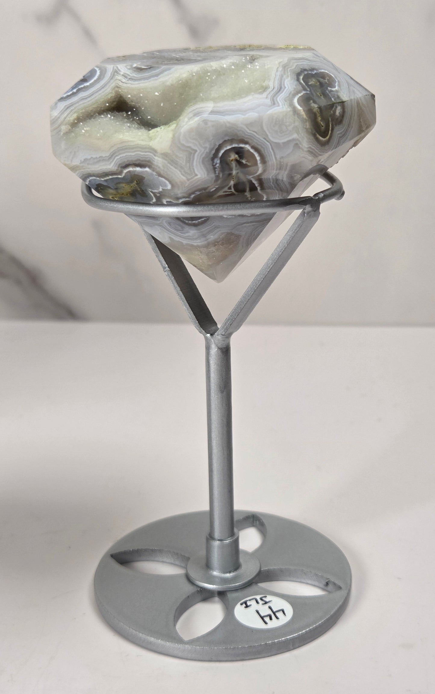 A polished agate gemstone with natural banding in white, gray, and green, displayed on a silver metal stand with a round base against a light marble-patterned background.