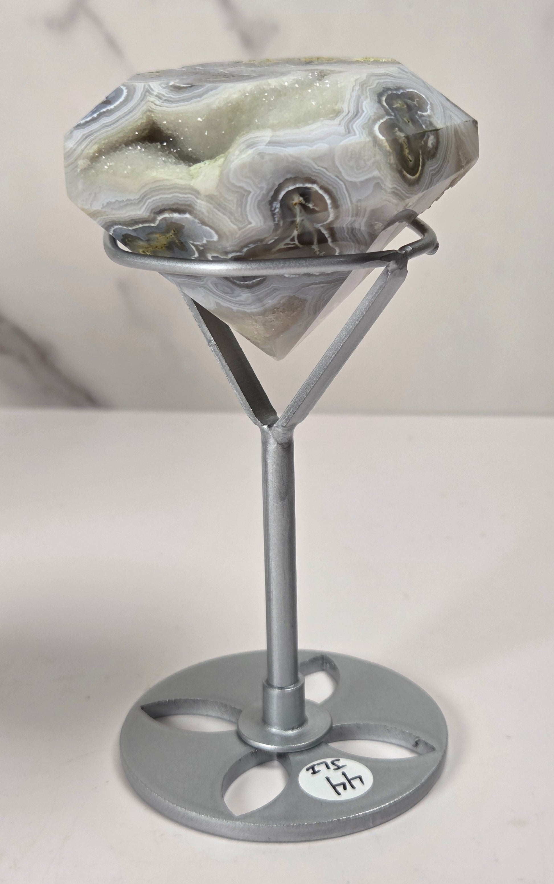 A polished agate gemstone with natural banding in white, gray, and green, displayed on a silver metal stand with a round base against a light marble-patterned background.
