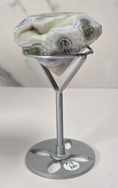 A polished agate gemstone with natural banding in white, gray, and green, displayed on a silver metal stand with a round base against a light marble-patterned background.