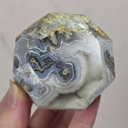 A hand holding a faceted octagonal agate crystal with banded gray, white, and black patterns and gold-colored mineral inclusions.