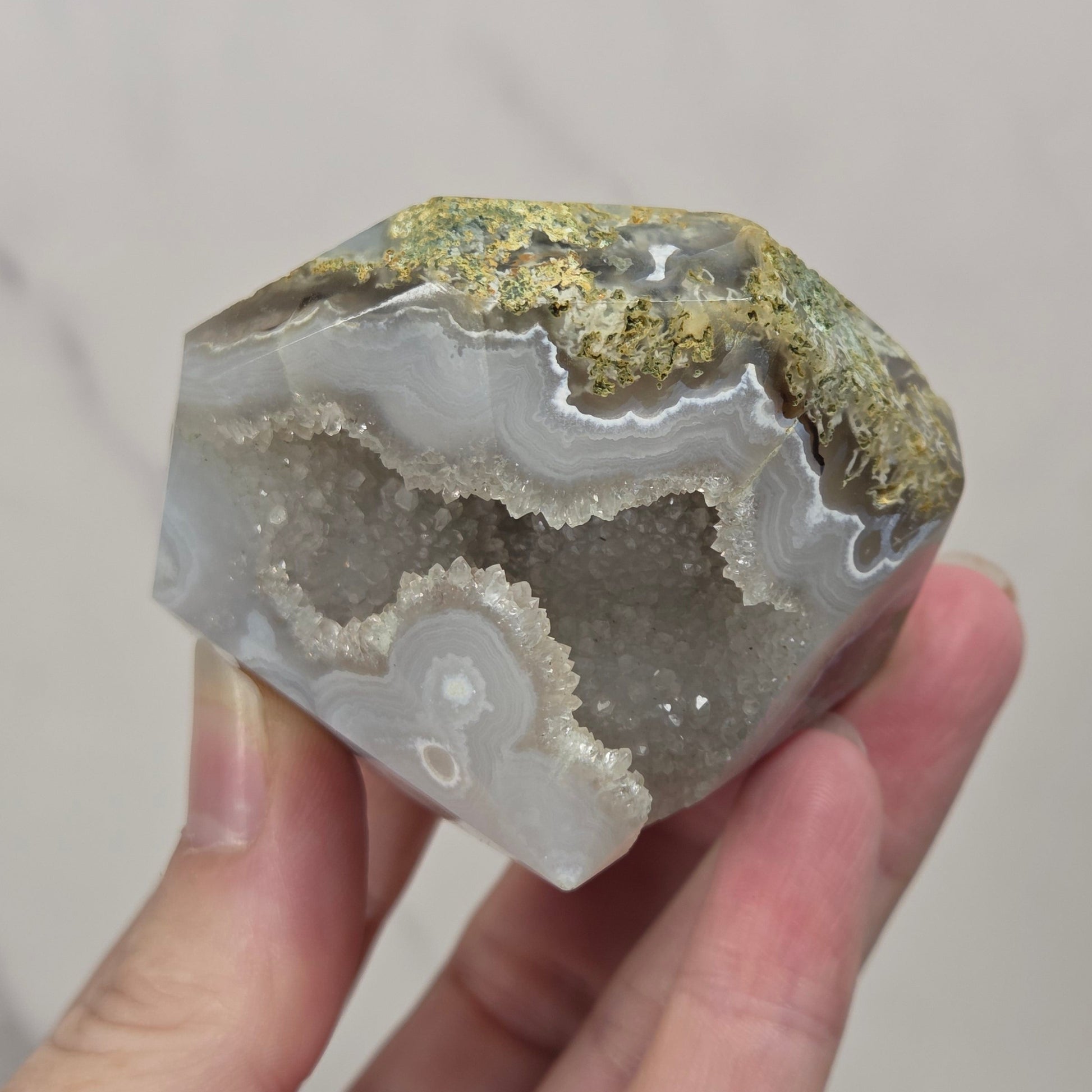 A hand holding a faceted geode specimen with layered white and gray agate bands and metallic gold crystalline deposits on top, set against a light marble-patterned background.