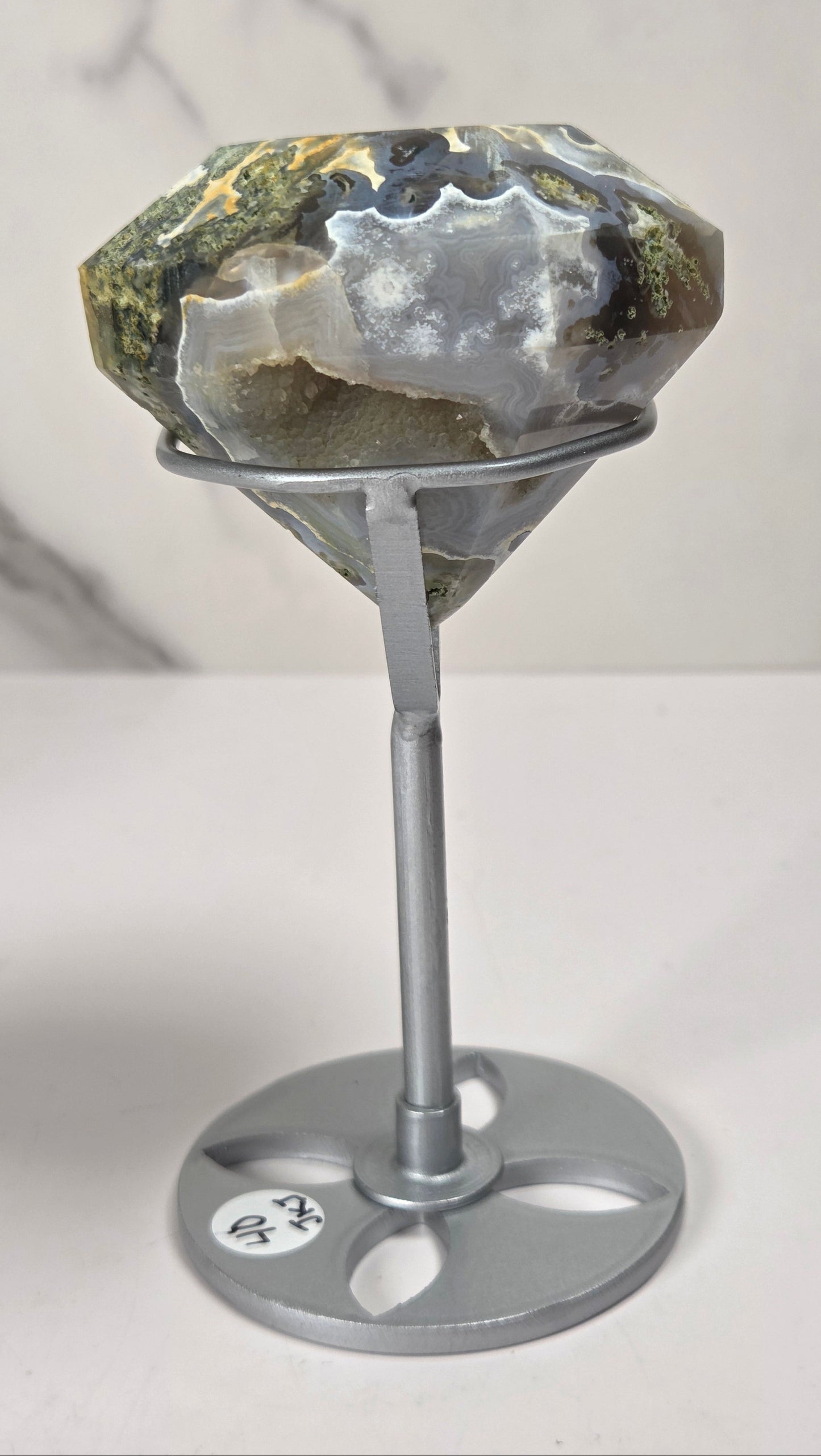 A large faceted gemstone with natural earthy tones of green, gray, and white, displayed on a silver metal stand against a light gray marble-patterned background.