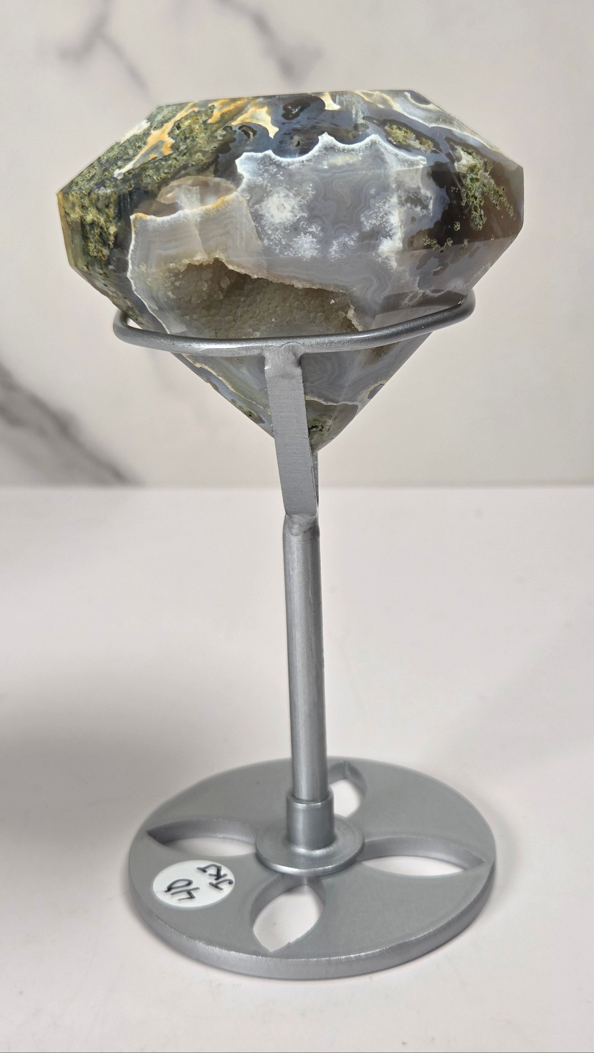 A large faceted gemstone with natural earthy tones of green, gray, and white, displayed on a silver metal stand against a light gray marble-patterned background.